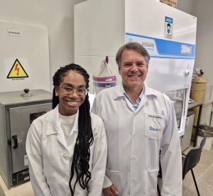 Anna e Carlos Henrique, pesquisadores, de pé lado a lado em um laboratório. Eles usam jaleco branco.