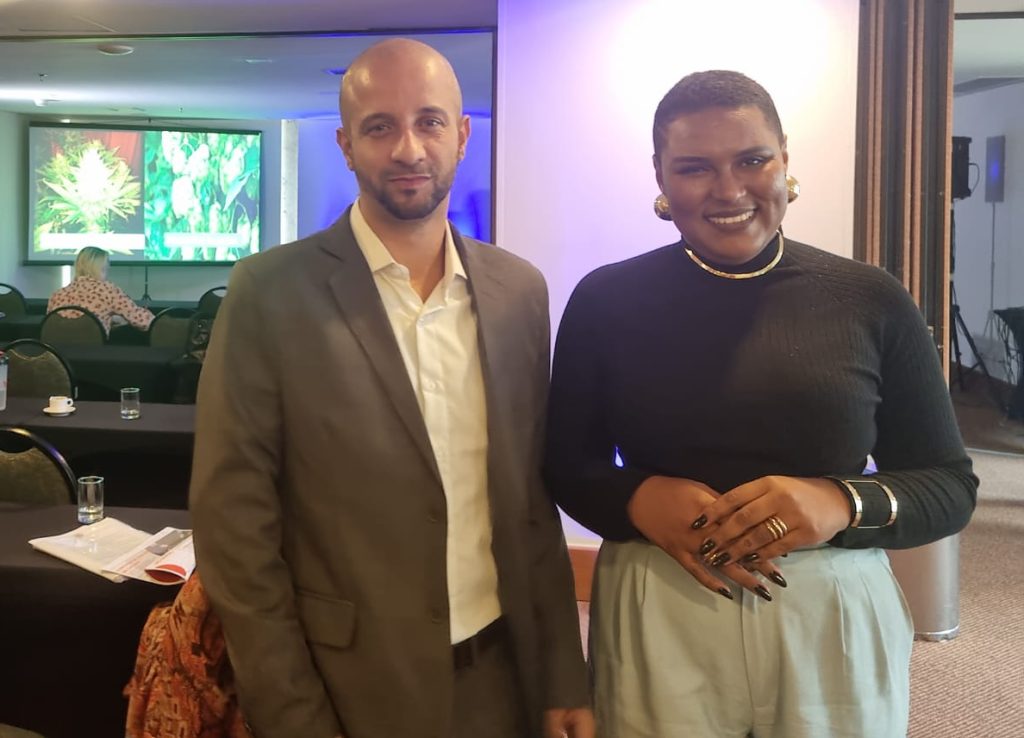 No Hemp Fair Experience Brasília, Thiago Ermano e Vitória Santos lado a lado. Ao fundo, pessoas assistem apresentação sobre Cannabis