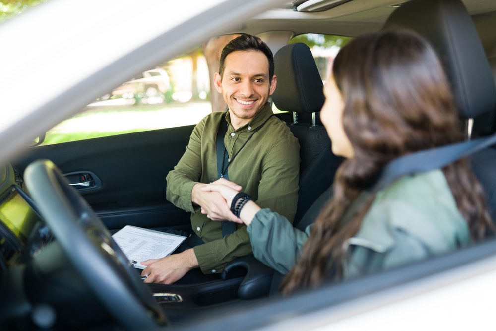 Instrutor de direção apertando a mão de aluna dentro de um carro de passeio, ambos usam cinto de segurança